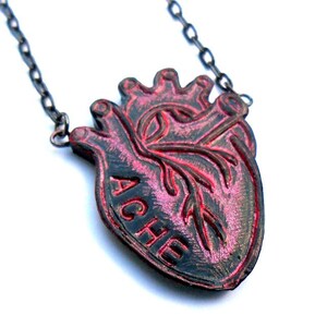 May include: A pendant necklace featuring a stylized anatomical heart design. The heart is black with red accents and the word "ACHE" is inscribed on the front. The necklace chain is a dark gunmetal color.