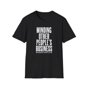 May include: Black t-shirt with white text that reads "MINDING OTHER PEOPLE'S BUSINESS" stacked vertically. Below the text is the hashtag "#ASSISTANTLIFE". The shirt is a crew neck style.