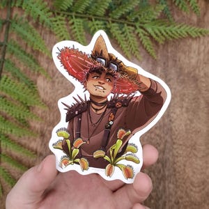 Puede incluir: Una pegatina con una ilustración estilizada de una persona con un sombrero marrón, detalles rojos y gafas. La persona lleva una camisa marrón y tiene atrapamoscas en primer plano. La pegatina está sujeta por una mano.