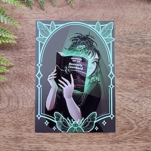 Könnte beinhalten: Illustration einer Person, die ein Buch mit dem Titel "Handbook for the Recently Deceased" liest. Das Kunstwerk zeigt eine dunkle Farbpalette mit neongrünen Akzenten, darunter stilisierte Schmetterlinge und geometrische Formen. Die Person trägt ein schwarzes Outfit.