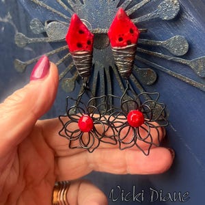 May include: Handmade earrings featuring red, black-dotted ceramic tops and wire-wrapped bases. Below, black wire flower designs hold red beads. The earrings are held in front of a blue background. The artist's signature, "Vicki Diane," is visible.