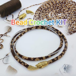 May include: A bead crochet kit with a necklace featuring brown, tan, and white beads. The necklace has a snake-shaped clasp. The kit includes a spool of thread, gold-colored metal findings, and a coiled strand of beads. The text "Bead Crochet KIT" is visible.