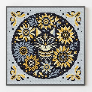 Puede incluir: Un bordado a punto de cruz enmarcado que presenta una cara de gato estilizada rodeada de girasoles y elementos florales. El diseño utiliza amarillo, azul y blanco sobre un fondo oscuro, con detalles decorativos en las esquinas.