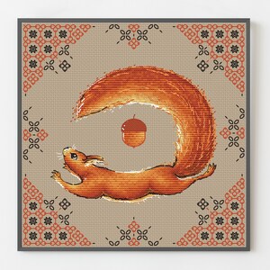 Può includere: Uno schema di punto croce con uno scoiattolo rosso con una coda folta, che tiene una ghianda nella sua bocca. Lo scoiattolo è circondato da un bordo decorativo di forme geometriche nere e arancioni.