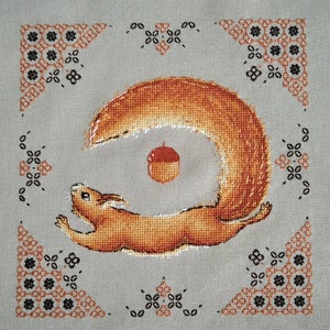 May include: A cross-stitch embroidery depicting a red squirrel with a bushy tail, leaping towards an acorn. The squirrel is framed by a decorative border of black and orange flowers.