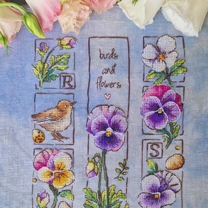 Może przedstawiać: Wzór haftu krzyżykowego przedstawiający ptaka, bratki i słowa "birds and flowers". Projekt zawiera brązowego ptaka, fioletowe i żółte bratki oraz litery "R" i "S". Tło to jasnoniebieska tkanina z różowymi i białymi kwiatami u góry.