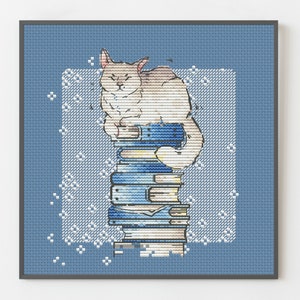 Könnte beinhalten: Kreuzstich-Kunstdruck einer Katze, die auf einem Stapel Bücher ruht. Die Katze ist hellbraun und weiß, mit einem mürrischen Gesichtsausdruck. Die Bücher sind blau und weiß, vor einem blauen Hintergrund mit weißen Akzenten, alles in einem dunkelgrauen Rahmen.