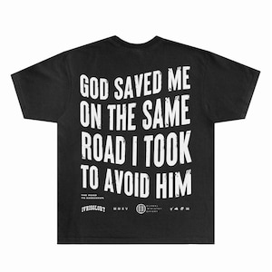 May include: Black t-shirt with white text: "GOD SAVED ME ON THE SAME ROAD I TOOK TO AVOID HIM." Below the text are the words "THE ROAD TO DAMASCUS" and "IVHISGLORY MMXV". A globe graphic and other text are also present.