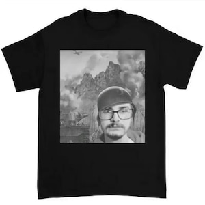 May include: Black t-shirt featuring a grayscale image of a person wearing glasses and a hat, superimposed over a war scene with mountains, tanks, and an airplane. The shirt is made of a soft, comfortable material.