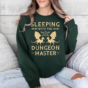 May include: A dark green sweatshirt with the text "Sleeping with the Dungeon Master" in yellow. Two yellow dragons are flanking a green and yellow dice.