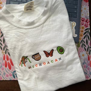 May include: White t-shirt with embroidered images of a traditional Mexican hat, a clay pot, a butterfly, and an avocado. The text "MICHOACAN" is embroidered below the images.