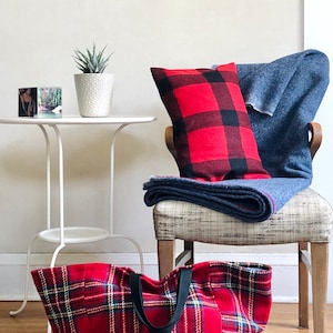 Puede incluir: Una bolsa de mano de lona roja y negra a cuadros con asas negras. La bolsa está sobre un suelo de madera con una alfombra debajo. En el fondo se ve una silla con un cojín rojo y negro a cuadros y una manta azul.