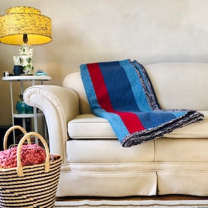 Könnte beinhalten: Eine blaue Decke mit rotem Streifen liegt auf einem cremefarbenen Sofa. Die Decke hat ein Streifenmuster und einen Fransenrand. Ein geflochtener Korb mit Streifenmuster und Griff steht auf dem Boden.