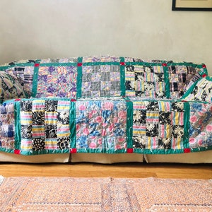 Puede incluir: Un edredón de retazos sobre un sofá. El edredón presenta una variedad de cuadrados estampados en tonos de azul, rosa, negro y blanco, enmarcados por un borde verde. Cuadrados rojos acentúan los bordes. El sofá está sobre un suelo de madera.