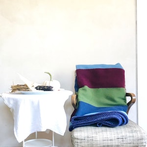 May include: A colorful striped blanket in shades of blue, burgundy, and green is draped over a chair. A small white table holds a white pumpkin, dried corn, and a plate. The scene has a rustic, autumnal feel.