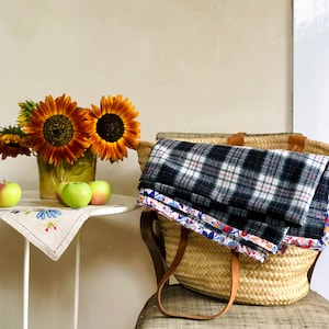 Könnte beinhalten: Ein Weidenkorb mit einer karierten Decke und einem Blumenstoff im Inneren. Der Korb steht auf einem Stuhl mit einem weißen Tisch davor. Auf dem Tisch stehen drei Äpfel und eine Vase mit Sonnenblumen.