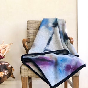 Könnte beinhalten: Eine graue Fleece-Decke mit einem abstrakten Design in Blau und Lila. Die Decke hat einen schwarzen Samtbesatz und liegt auf einem Holzstuhl.