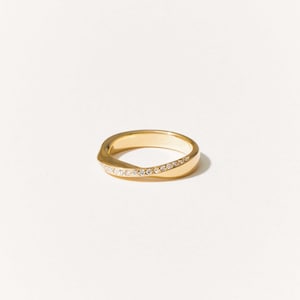 Puede incluir: Un anillo dorado con un diseño curvo. El anillo presenta una fila de pequeñas piedras preciosas transparentes engastadas en un lado. El anillo se encuentra sobre un fondo blanco liso, resaltando su elegancia.