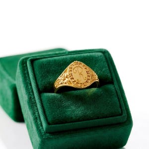 May include: A gold signet ring featuring an oval face engraved with the cursive letter R, surrounded by a beaded border and intricate floral detailing along the band, presented inside a deep green velvet ring box.