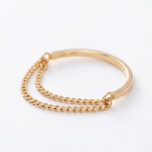 May include: A gold ring featuring a smooth, solid band that transitions into a delicate, double-layered chain section. The chain is attached to both ends of the solid band, creating a continuous circular shape with a polished, metallic finish.