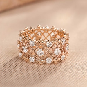 Puede incluir: Un anillo de oro rosa con un diseño de celosía ancho e intrincado con un acabado texturizado. La estructura de metal está adornada con múltiples piedras redondas, claras y facetadas engastadas en las intersecciones del patrón entrecruzado.