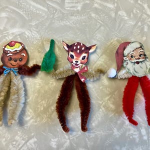 以下が含まれることがあります： 手作りのクリスマスオーナメント3つ。 ロリポップを持ったジンジャーブレッドマン、トナカイ、サンタクロース。 各フィギュアは、モールと紙の切り抜きで作られており、お祝いの色とディテールが施されています。