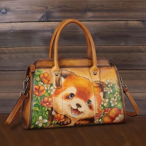 May include: A tan leather handbag featuring a hand-painted illustration of a red panda surrounded by orange and white flowers. The bag includes two top handles, a detachable shoulder strap, and metal hardware attachments on the sides.