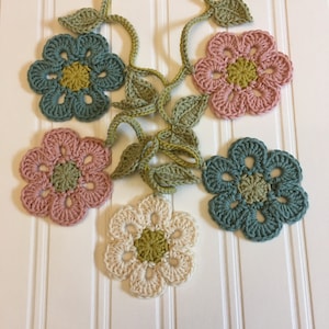 May include: A garland of crocheted flowers in shades of green, pink and cream. The flowers are connected by crocheted leaves.