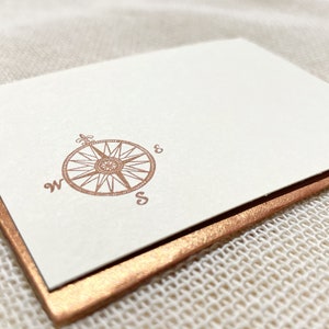 May include: A white card with a copper colored border. The card has a copper colored compass design with the letters "W" and "S" printed on the card.