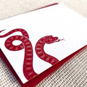 May include: A red snake with a patterned design is printed on a white card with a red border. The card is blank and can be used for writing a note.
