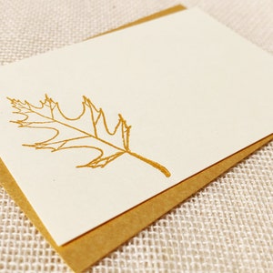 May include: A blank card with a gold foil oak leaf design. The card is folded in half and has a brown envelope.