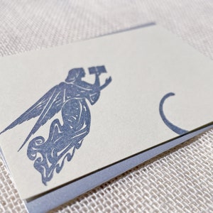May include: A white card with a blue angel design. The angel is holding a trumpet and has wings. The card has a blue border.