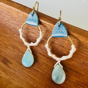 Op de afbeelding: Een paar hangende oorbellen met een driehoekige bovenkant van metallic lichtblauw leer, een goudkleurige cirkelvormige ring omwikkeld met off-white koord en een gladde, druppelvormige lichtblauwe steen onderaan elke ring.