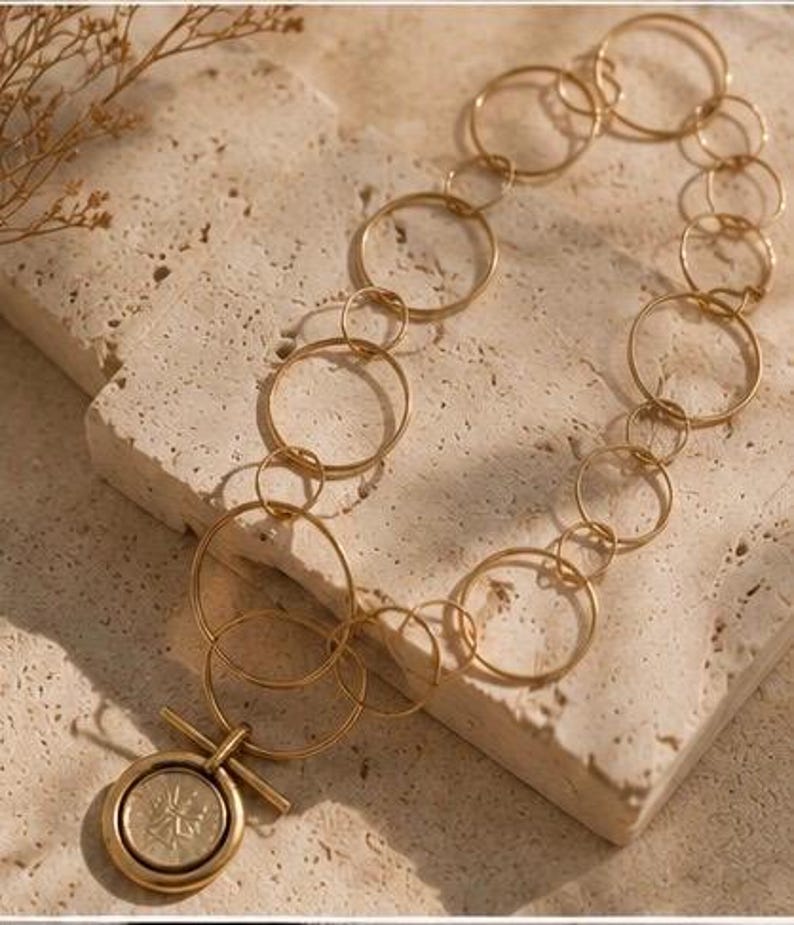 May include: A gold-toned necklace featuring a series of interconnected circular links of varying sizes. The chain leads to a circular pendant with a central embossed coin-like design, secured by a horizontal bar toggle clasp mechanism.
