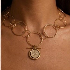May include: A gold-toned necklace featuring a series of interconnected circular links of varying sizes. The centrepiece is a circular pendant with a horizontal bar detail, containing a textured coin-like element with an embossed profile design.
