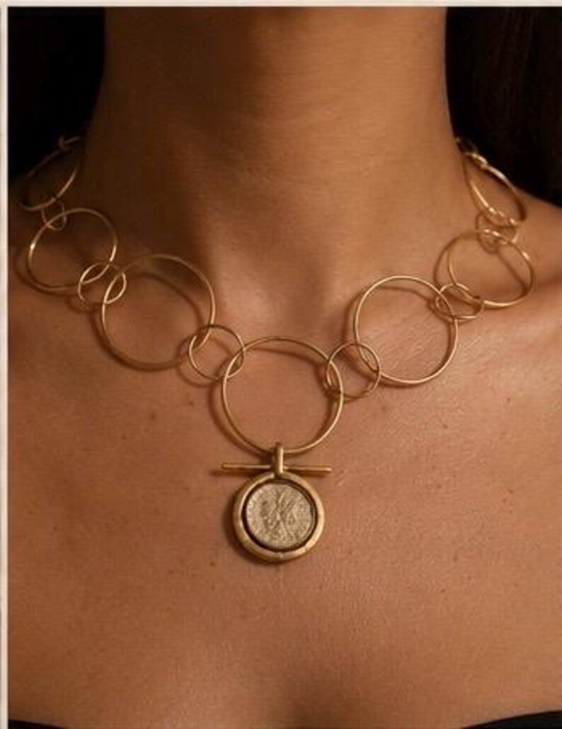 May include: A gold-toned necklace featuring a series of interconnected circular links of varying sizes. The centrepiece is a circular pendant with a horizontal bar detail, containing a textured coin-like element with an embossed profile design.