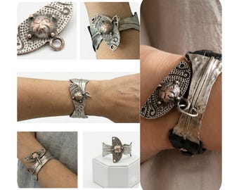 May include: A silver-toned cuff bracelet featuring a textured, semi-circular metal plate with a central raised dome and swirling patterns. The bracelet includes a dark, possibly leather or fabric, band and a small attached metal ring charm.