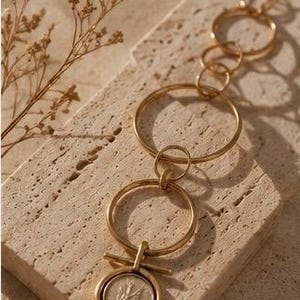 May include: A gold-toned necklace featuring a series of interlocking circular links of varying sizes. The necklace includes a central pendant consisting of a circular coin-like charm suspended from a horizontal bar attached to a larger ring.