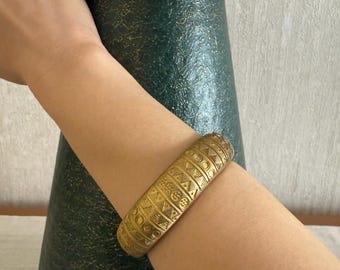 May include: A gold-coloured, patterned bangle bracelet. The bracelet is thick and features geometric designs. The bracelet is worn on an arm, with a dark green and gold decorative jar in the background.