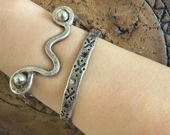 May include: Two silver bracelets are shown on a wrist. One is a wavy design with ball accents, and the other is a cuff with a repeating pattern of crosses and starbursts. The bracelets have a vintage, handcrafted appearance.