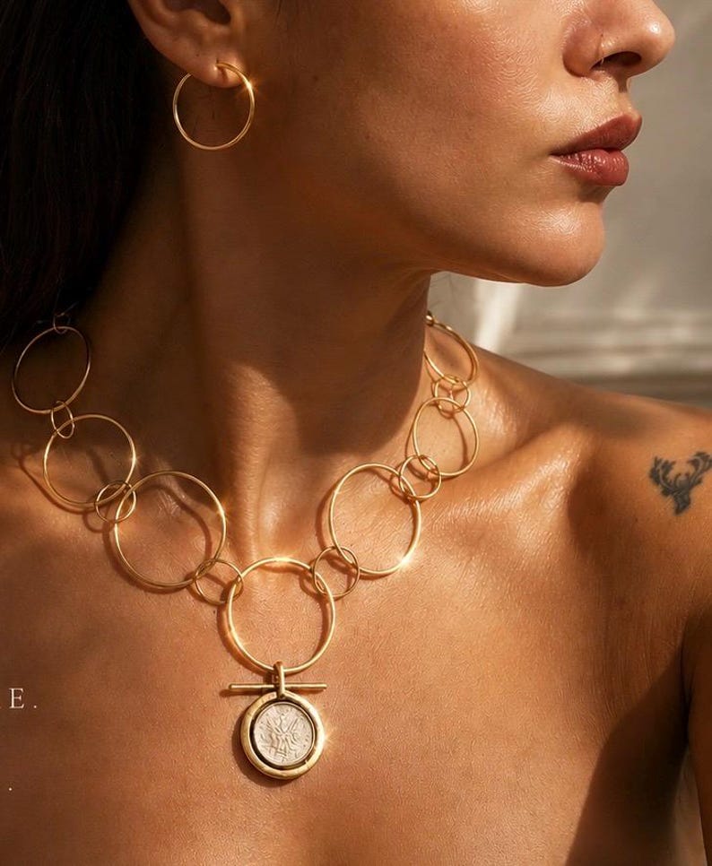 May include: A gold-toned necklace featuring a series of interconnected circular links of varying sizes, with a central pendant consisting of a circular frame holding a silver-coloured coin-like disc, complemented by a matching simple gold hoop earring.