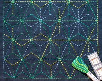 May include: A dark blue fabric square with a geometric pattern stitched in green, yellow, and blue thread. The fabric is surrounded by a white border. A spool of green and yellow thread is in the lower right corner of the image. A small wooden box with Japanese text is also in the lower right corner.