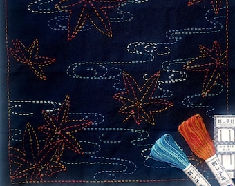 May include: A dark blue fabric square with a Japanese sashiko embroidery design of stylized maple leaves and clouds. The fabric is surrounded by three spools of embroidery floss in shades of blue, orange, and red.
