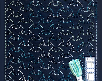 May include: A blue fabric square with a white and green geometric pattern stitched on it. A spool of green embroidery floss and a package of needles are on the right side of the fabric.
