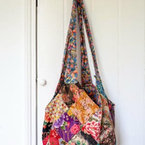 Op de afbeelding: Een patchwork tote bag met verschillende bloemenpatronen in tinten rood, roze, oranje, geel, blauw, groen en paars. De tas hangt aan een witte deur met zwarte hartvormige haken.