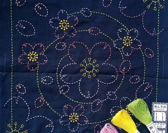 May include: A dark blue fabric square with a design of pink, yellow, and green flowers embroidered on it. The fabric is surrounded by a white border. There are three spools of embroidery floss in pink, yellow, and green.