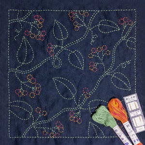 May include: A dark blue fabric with a design of leaves and berries stitched in green, orange, and pink thread. The fabric is surrounded by a white border. There are three spools of thread in the bottom right corner of the image, one green, one orange, and one pink. The spools are labelled in Japanese.