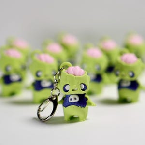 May include: A collection of small, green zombie figurines with pink brains and blue shirts. One figurine is attached to a silver keychain. The figurines are arranged on a white surface, with several in focus and others blurred in the background.
