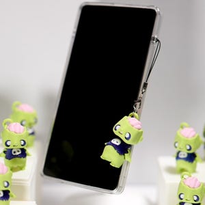 May include: A smartphone with a clear case and a zombie charm attached. The charm is lime green with a pink brain, a blue shirt, and a skull design. Several zombie charms are arranged on white platforms.