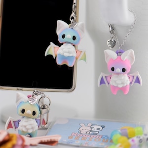 May include: Four bat-shaped charms featuring pastel-colored bodies with white chest fur, large dark eyes, and small wings. Each charm is attached to a silver-toned metal chain or clasp, with one charm featuring a small heart-shaped tag labeled Made with Love.